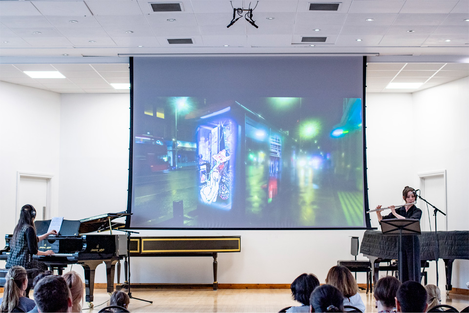 Two students performing on a piano and violin, one of whom composed the song, on a stage with an audience watching them.